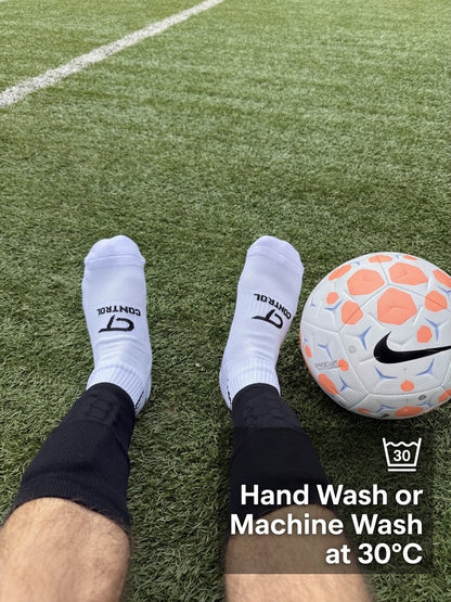 White athletic socks with black PlayStation-style logos worn on feet, shown on a green turf field next to a white soccer ball with orange and blue accents. Text overlay reads 'Hand Wash or Machine Wash at 30°C' with a wash icon.